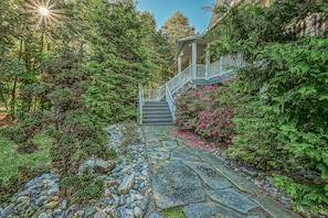 A stone pathway leading to a house with a porch and stairs, surrounded by dense greenery and ferns.