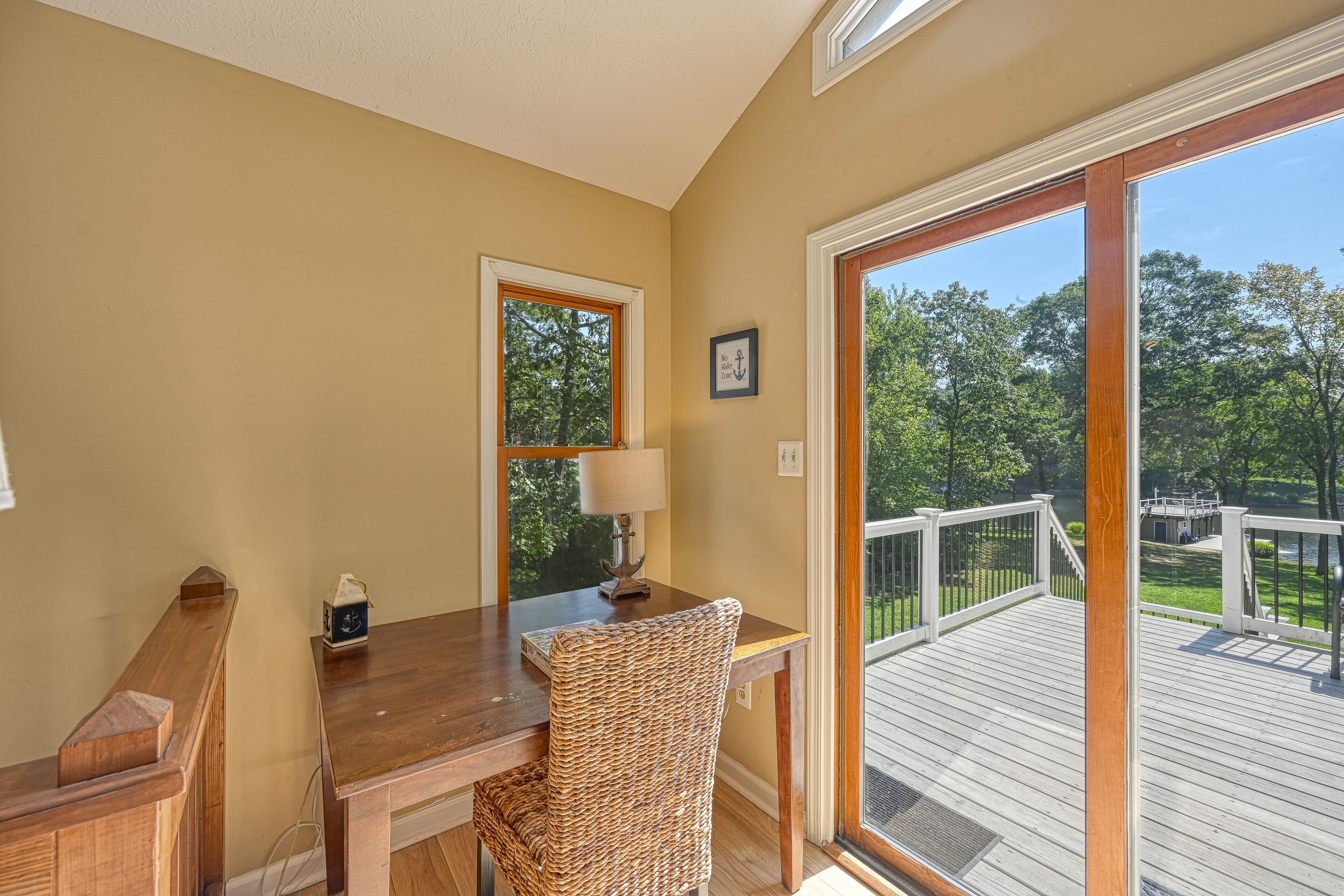 Une pièce avec un bureau en bois, une chaise en osier et une vue sur une terrasse et des arbres, visible à travers une grande porte vitrée.