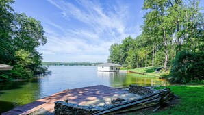 Terraza junto al lago con pared de piedra y tablones de madera, rodeada de abundante vegetación y cielo despejado.