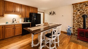 Une cuisine avec des meubles en bois, un réfrigérateur noir, une table à manger avec des chaises blanches et une cheminée en pierre.