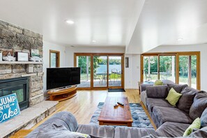 A spacious living room with a stone fireplace, a large flat-screen TV, a grey sofa, and a wooden coffee table. - Lakefront home with two kitchens, a dock, firepit (Bumpass)