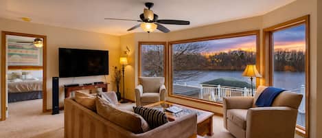 Una sala de estar con sofá, sillas, televisor y vista a un lago al atardecer.