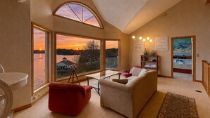 A spacious living room with a large window overlooking a lake at sunset, a red armchair, a beige sofa, and a dining area.