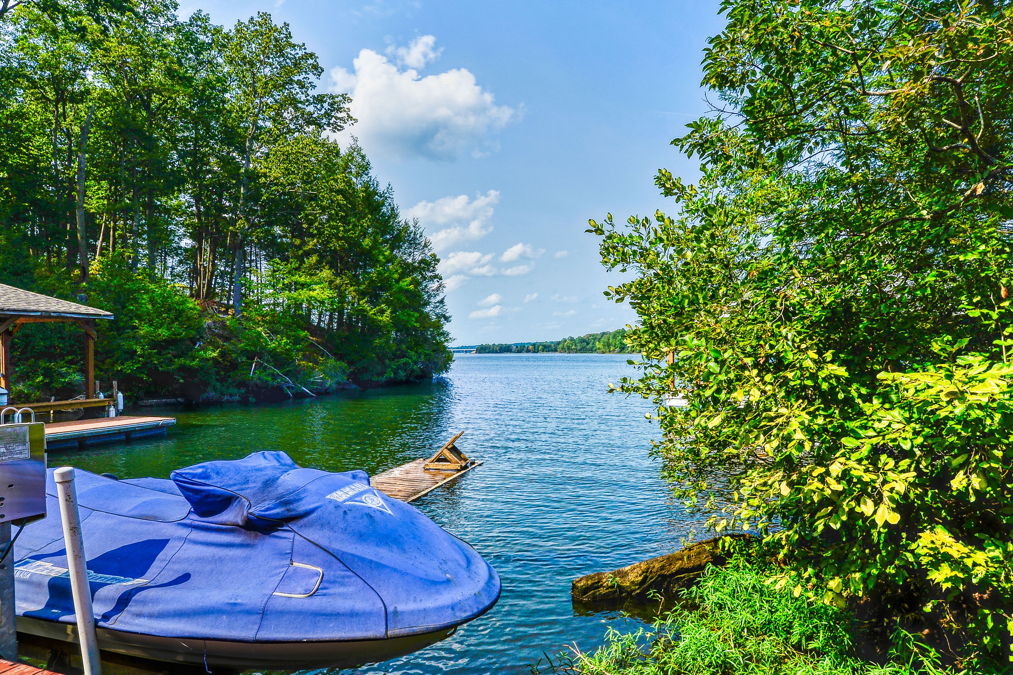 A dock with a blue inflatable boat and a wooden boat on calm water surrounded by lush greenery.