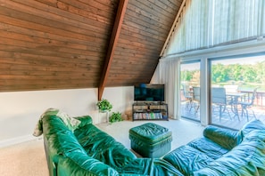 A modern living room with a sloped ceiling, a large green sofa, a television, and a view of an outdoor deck.