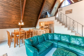 A spacious living area with a green leather sofa, wooden dining table, and chairs, a kitchen in the background, and a staircase with decorative railings.