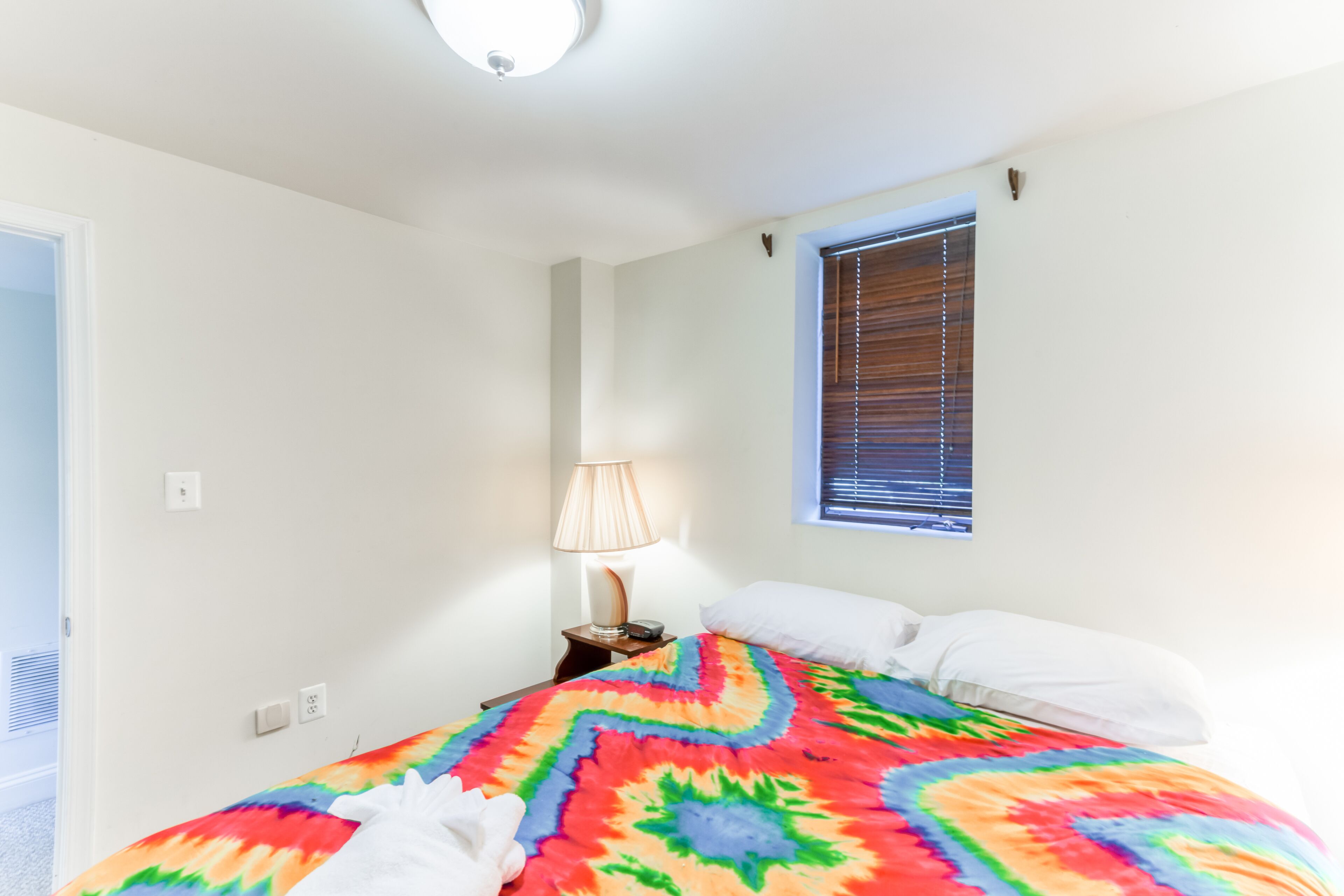 Un dormitorio con una colcha colorida, una lámpara de noche, una ventana con persianas y una puerta que da a otra habitación.