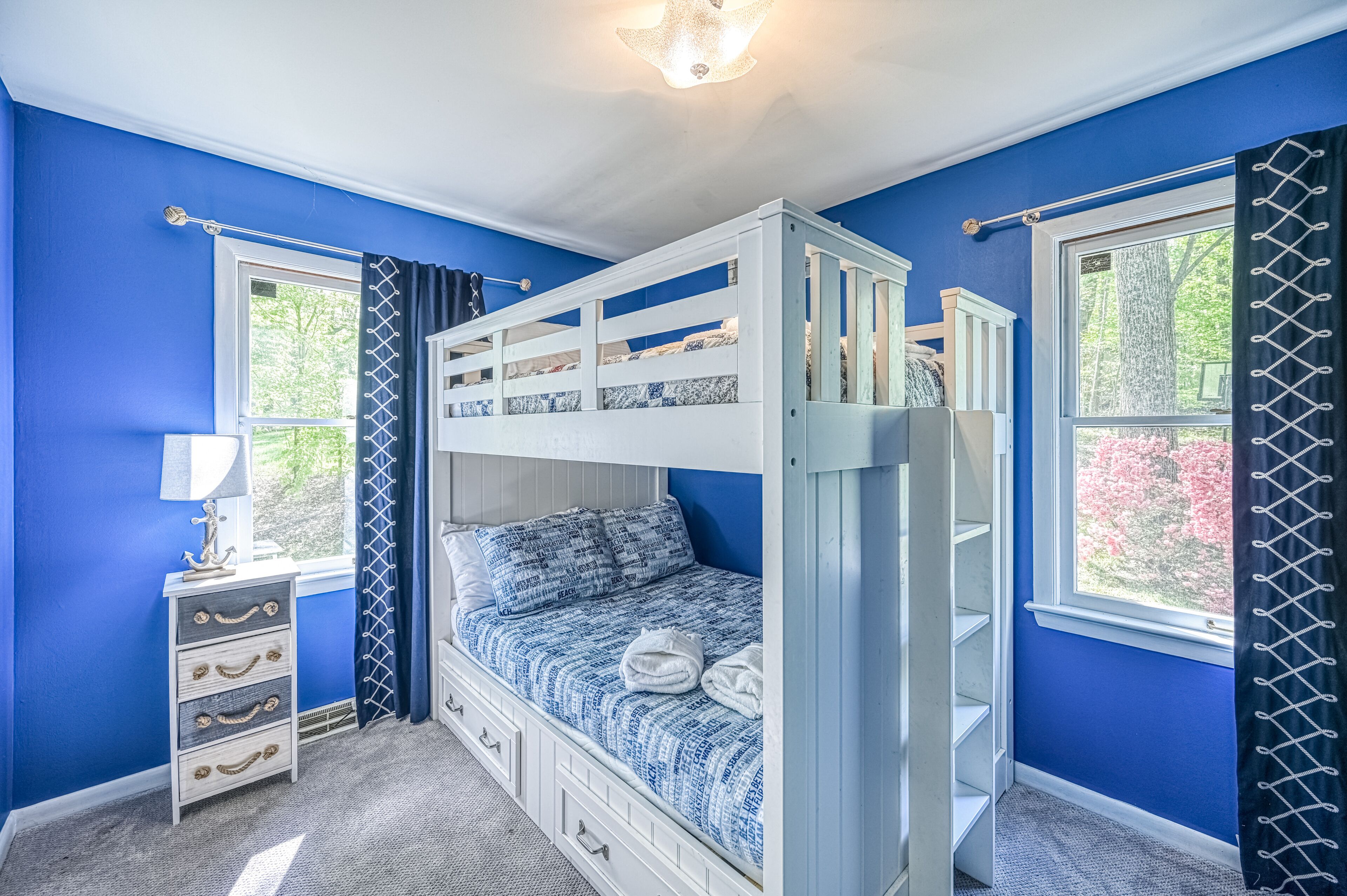 Habitación con literas, una pared azul, ventana con vista a árboles, cómoda con cajones y una lámpara.
