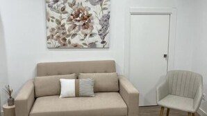 A living room with a sofa, two round wooden tables, a chair, a floral painting, and an air conditioning unit. - APARTAMENTOS SAN MIGUEL (Jerez de los Caballeros)