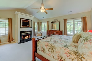 A bedroom with a fireplace, a bed with floral bedding, a ceiling fan, and large windows with curtains.