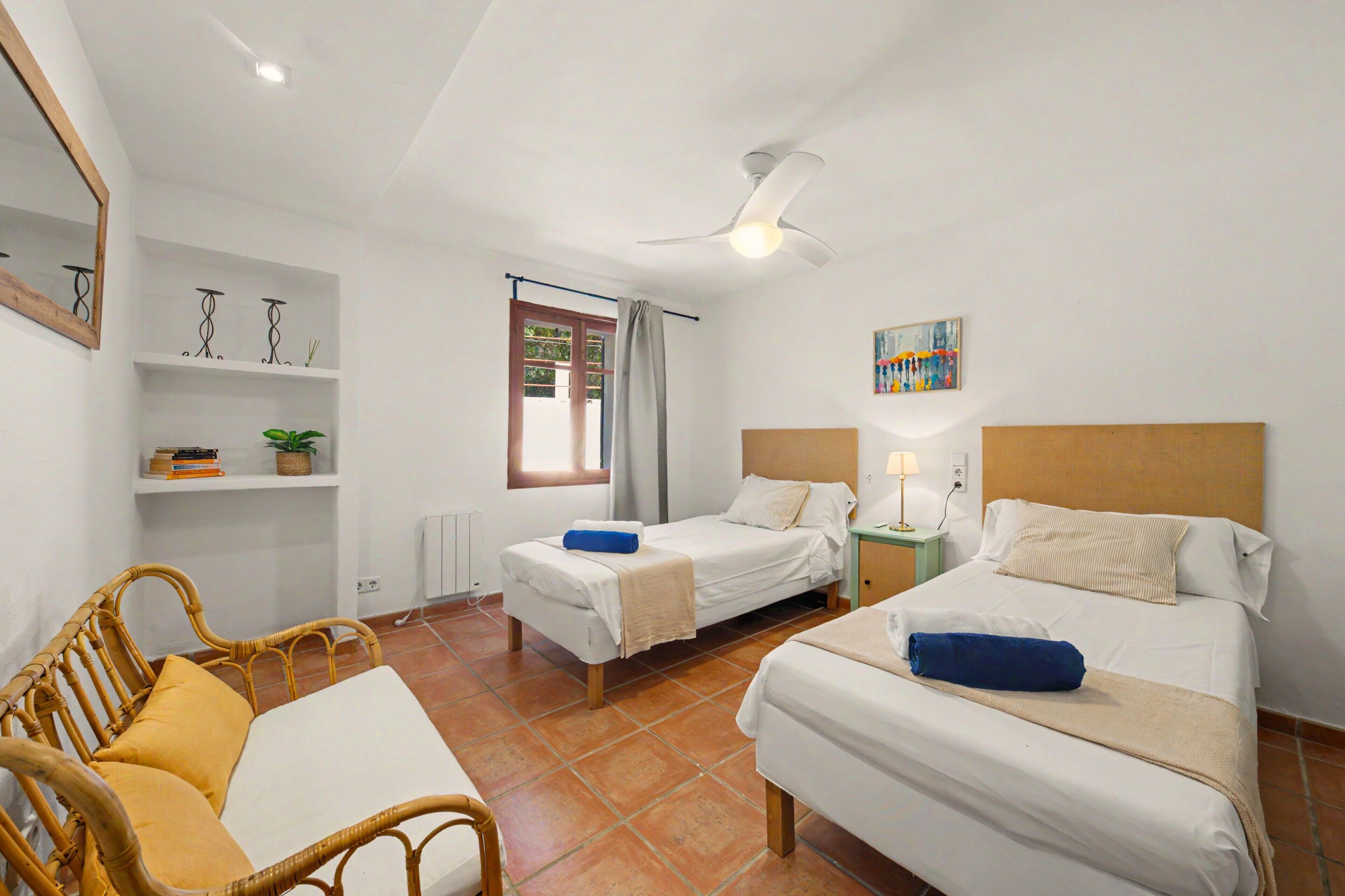A hotel room with two beds, a chair, a small table, a ceiling fan, and a window with curtains.