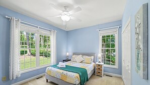 Ein Schlafzimmer mit einem Bett, zwei Nachttischen, einem Deckenventilator und großen Fenstern mit Vorhängen.