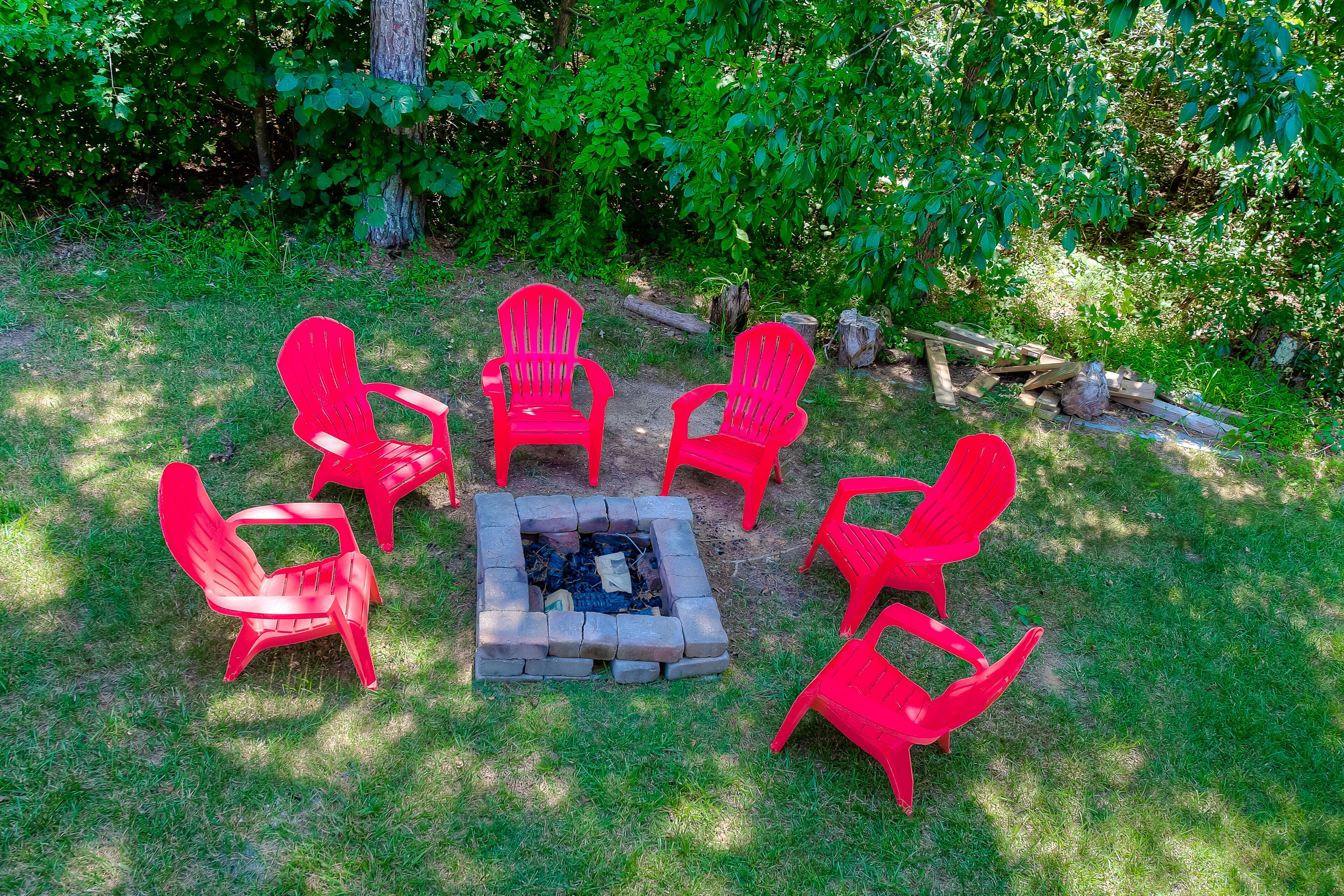 A fire pit surrounded by six red Adirondack chairs in a grassy outdoor area with trees in the background.