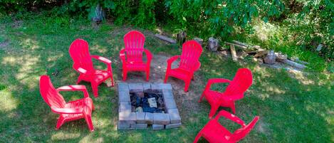 A fire pit surrounded by six red Adirondack chairs in a grassy outdoor area with trees in the background.
