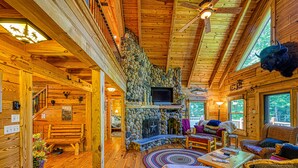 A log cabin interior with a stone fireplace, wooden furniture, and a television.