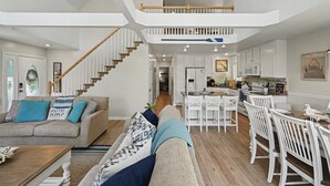 Een ruime woonkamer met een hoge plafond, een trap, een keuken met witte kasten en een eethoek met witte stoelen.