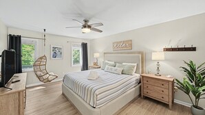 Een slaapkamer met een bed, een plafondventilator, een nachtkastje met een lamp, een houten dressoir, een stoel en een kamerplant.