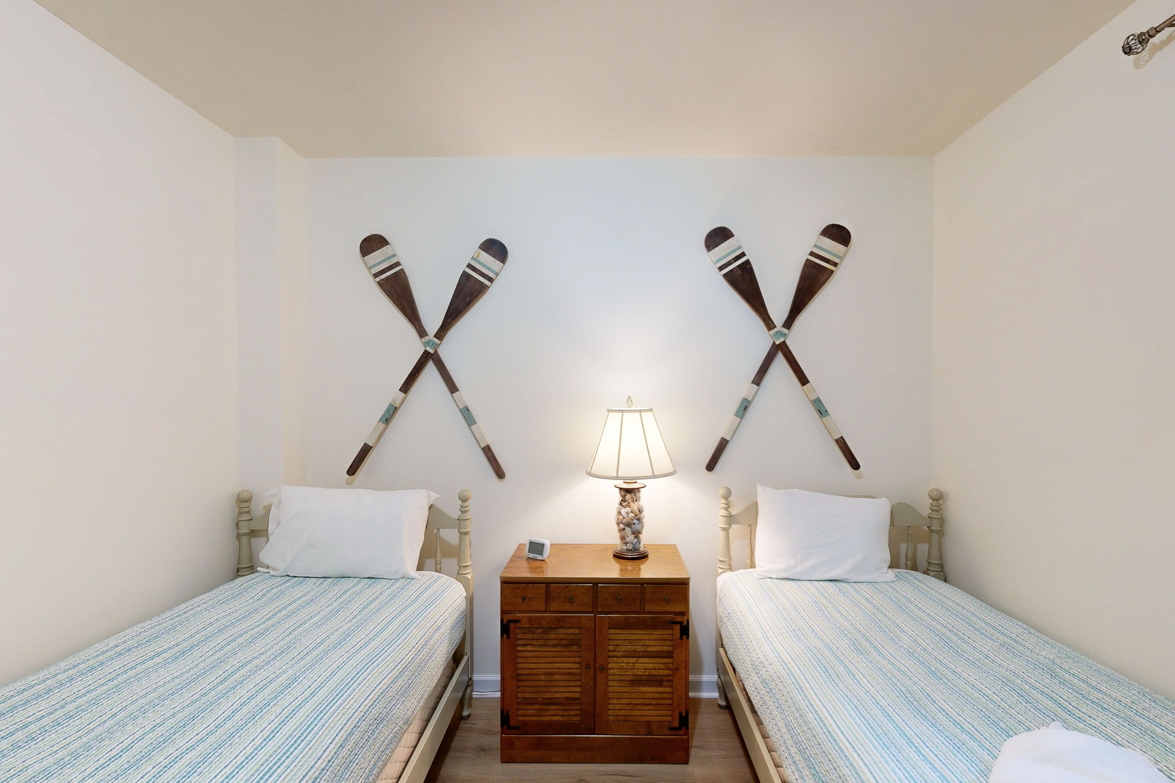 A bedroom with two beds, a wooden nightstand with a lamp, and two wooden paddles mounted on the wall.