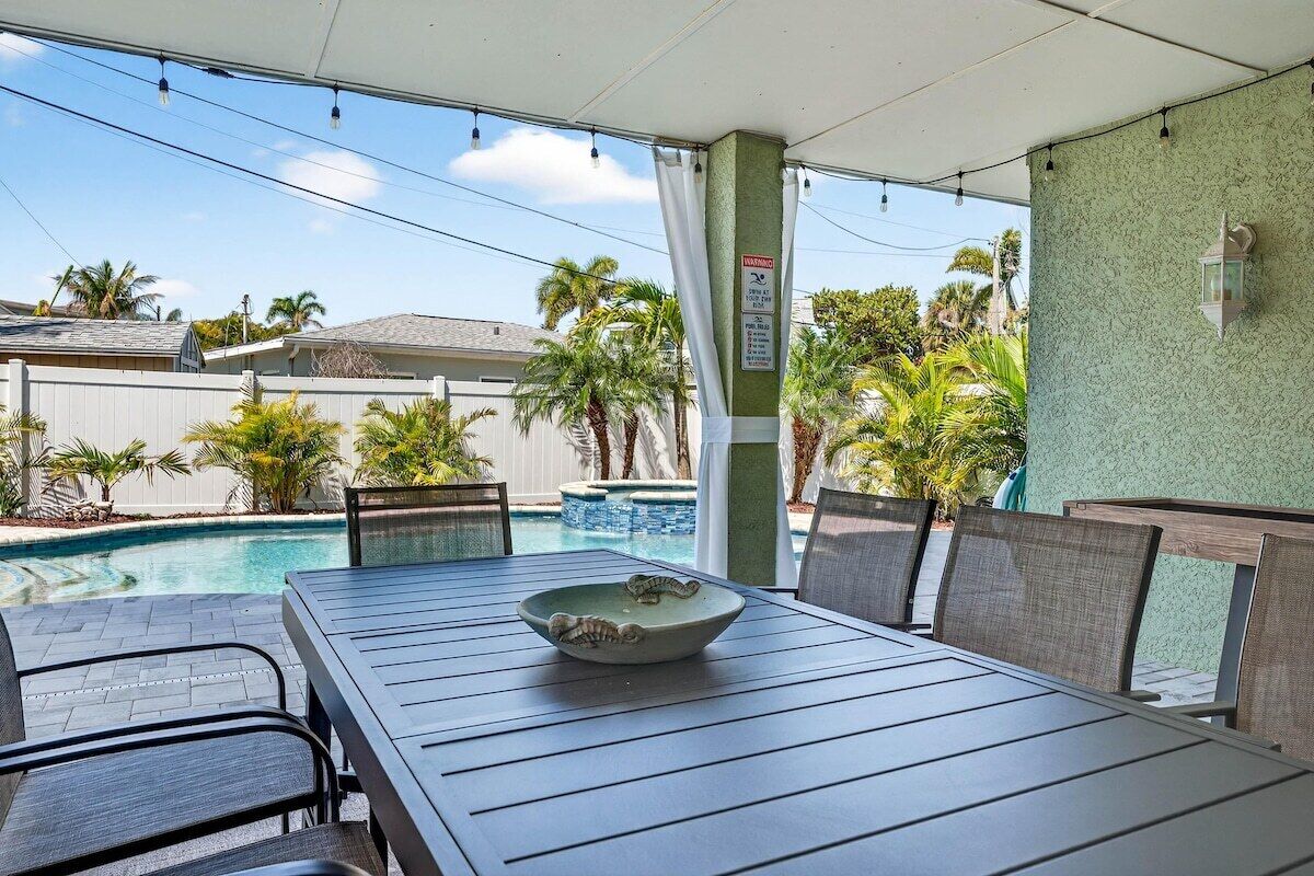 An outdoor patio with a table and chairs, a pool, and a view of a residential area with palm trees.