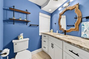 Ein Badezimmer mit blauer Wand, einer weißen Toilette, einem hölzernen Regal mit Pflanzen, einem Spiegel und einem Handtuch mit Blätchenmuster.