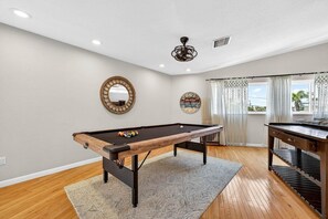 A pool table with balls, a mirror, and a ceiling fan in a room with wooden flooring and a window with curtains. - Blue Lagoon 3BR Pool Home Near the Beach (Redington Beach)