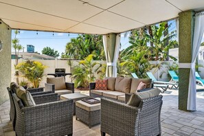Un espacio de estar techado al aire libre con mobiliario de mimbre y vistas a una piscina y palmeras.