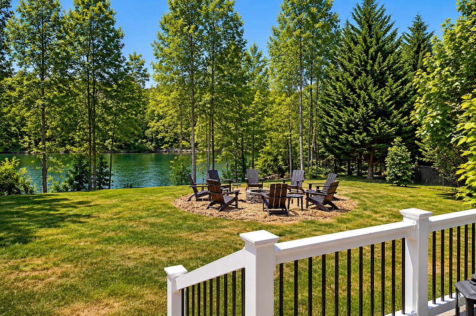 A deck with a white railing, surrounded by a grassy lawn and trees, with a fire pit and Adirondack chairs.