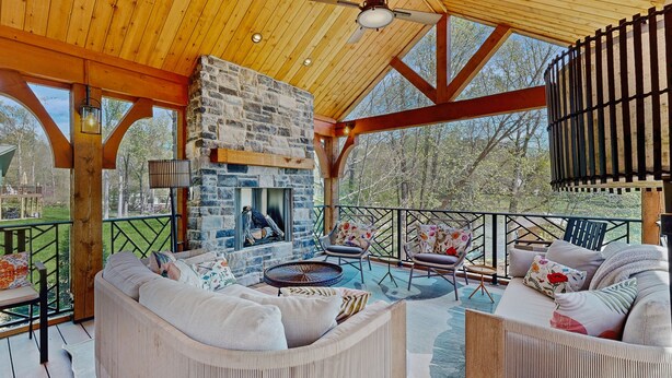 A covered outdoor seating area with wicker furniture, a stone fireplace, and a view of the surrounding trees. - Tranquil lakefront retreat with hot tub, screened porch, kayaks, and more (Bumpass)