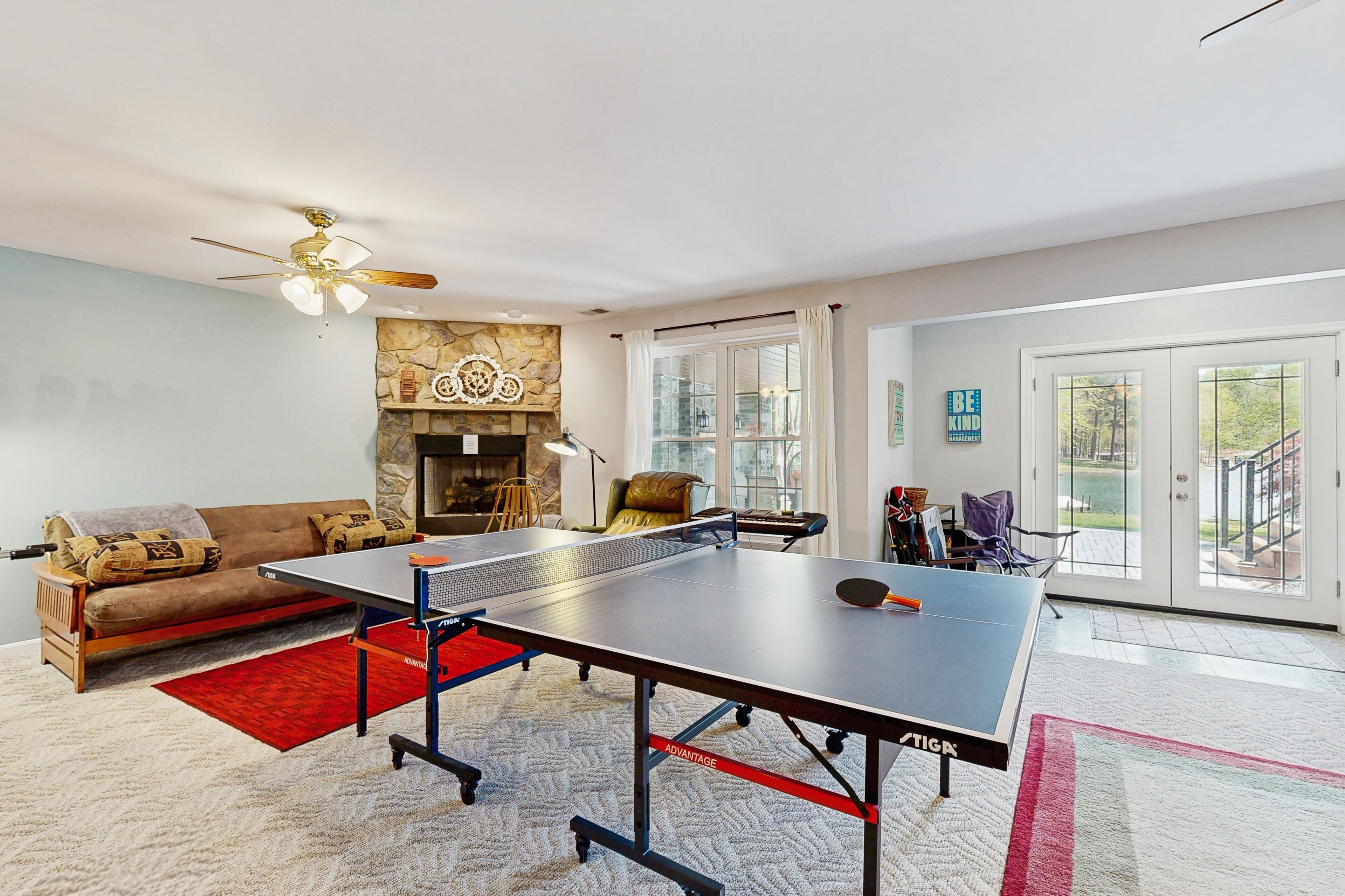 A spacious living room with a ping pong table, a sofa, a fireplace, and a large window offering a view of the outdoors.