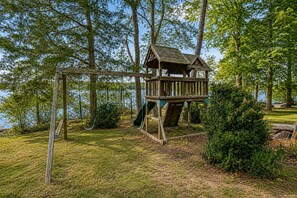 Ein hölzernes Spielhaus mit Rutsche, umgeben von Bäumen und Gras.