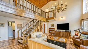 Ein geräumiges Wohnzimmer mit hoher Decke, einer Holztreppe, einem großen Kronleuchter, einem Fernseher und einem Schachbrett.