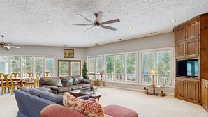 A spacious living room with a large sofa, a coffee table, and a television. There are multiple windows providing natural light and views of the outdoors. - Rustic lakehouse with views, kayaks, Wifi, Central AC & cozy firepit nights (Bumpass)