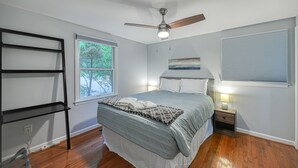 Un dormitorio con cama, ventilador de techo, ventana, mesita de noche y estante en la pared.