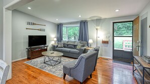 Una sala de estar con un sofá gris, una mesa de centro y un televisor de pantalla plana. Hay dos lámparas sobre mesas auxiliares, una ventana con cortinas y una puerta que da al exterior.