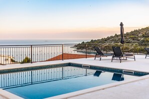 Ein Pool mit Liegestühlen und Sonnenschirm, mit Blick auf einen Strand und eine hügelige Landschaft.