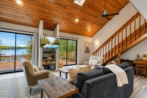 A spacious living area with a wooden ceiling, a large window overlooking a lake, a staircase, and a cozy seating arrangement.