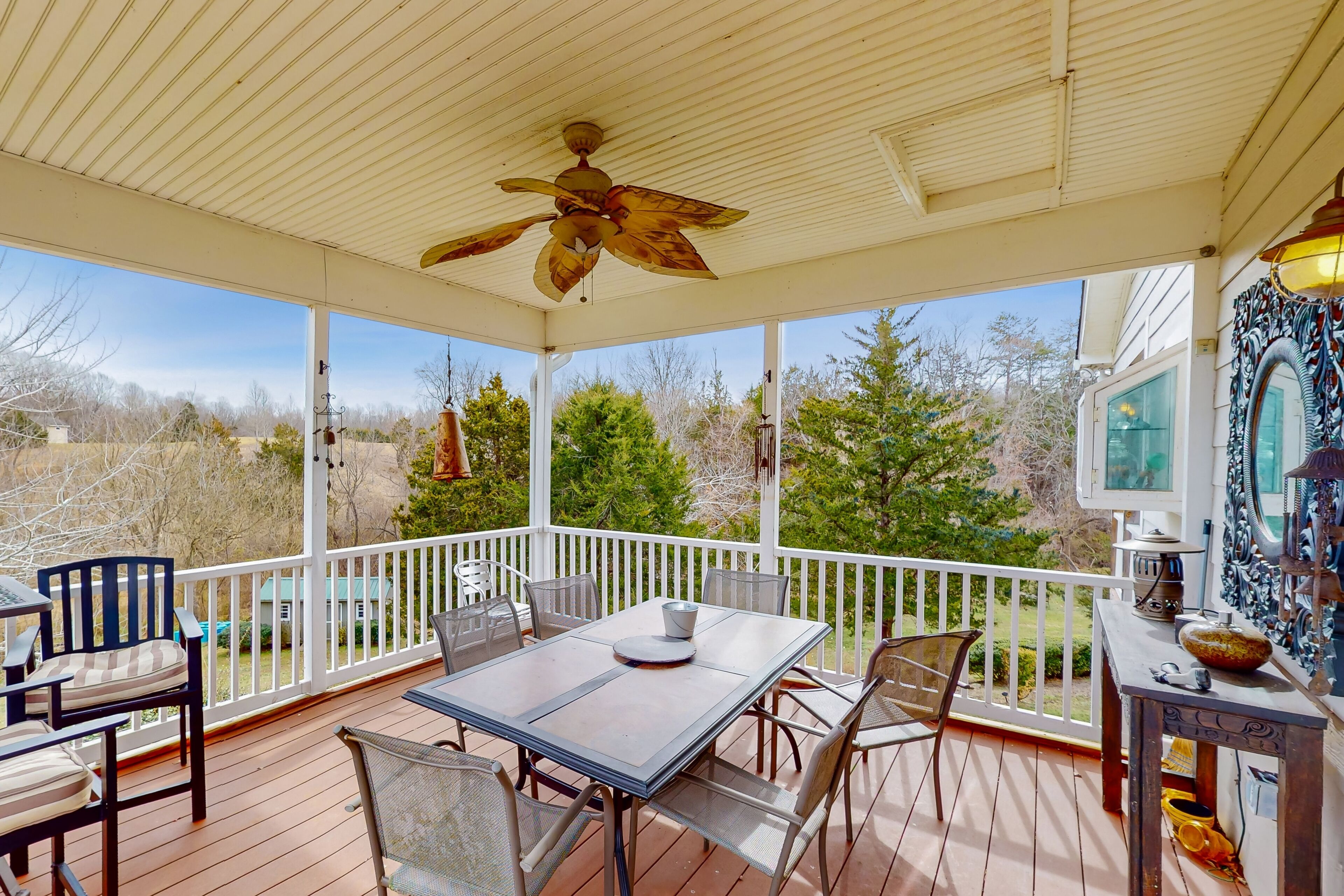Une terrasse extérieure couverte, dotée d’une table et de chaises, d’un ventilateur de plafond et offrant une vue sur le paysage environnant.