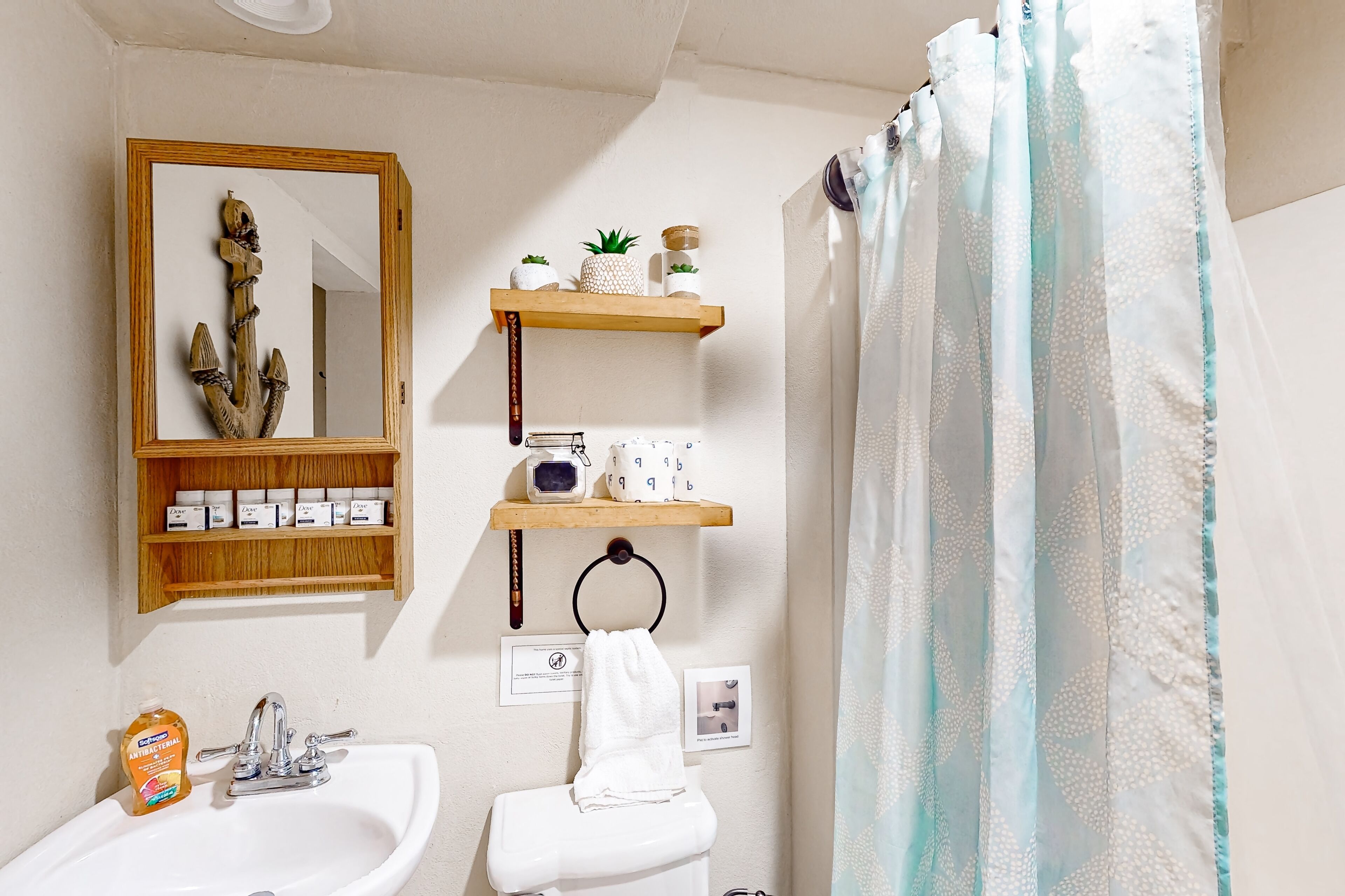 A bathroom with a white sink, a mirror, a toilet, and a shower curtain. There are wooden shelves above the toilet with toiletries and a small plant.