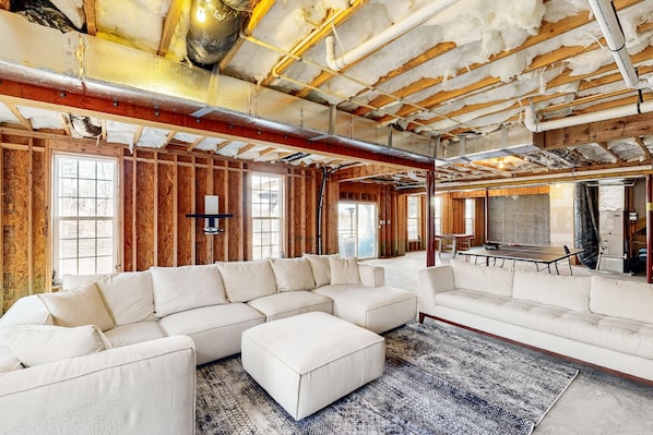 A spacious living room with a large sectional sofa, a small ottoman, and a patterned rug. The room has exposed wooden beams and a high ceiling with visible ductwork.