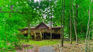 Eine Holzhütte mit einer überdachten Veranda und einer Treppe, die dazu hinauf führt, umgeben von dichtem Wald.