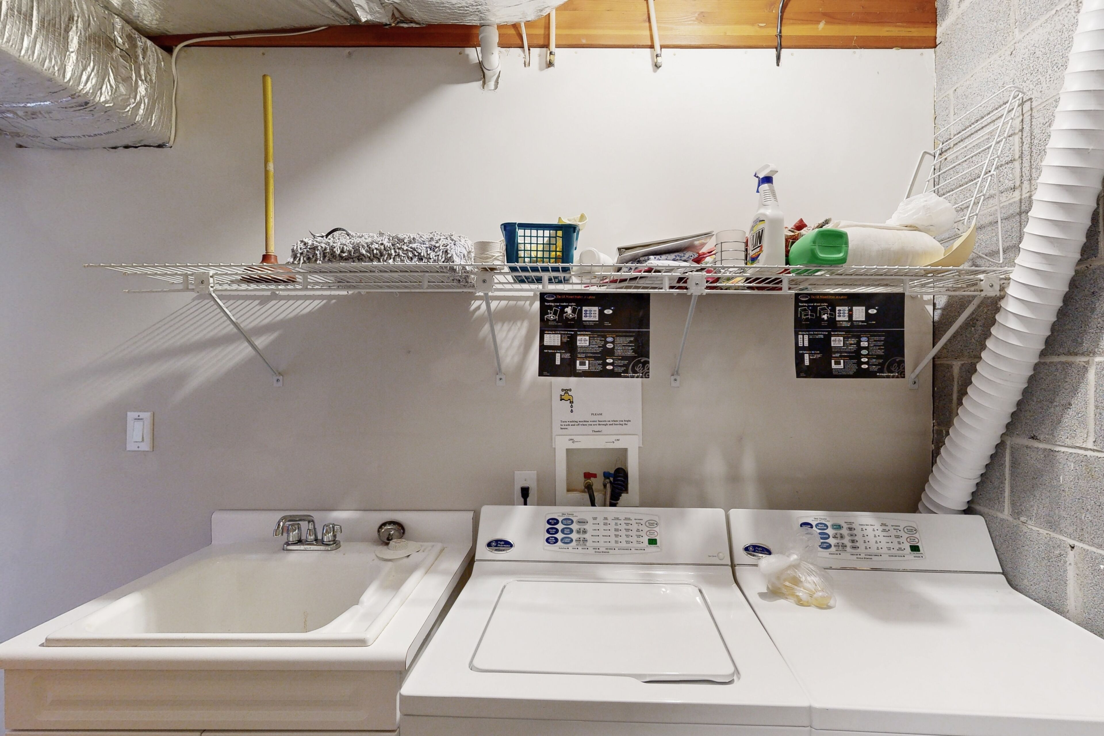 A laundry area with a washing machine, dryer, and a sink. There are shelves above holding various items like bottles and a basket.
