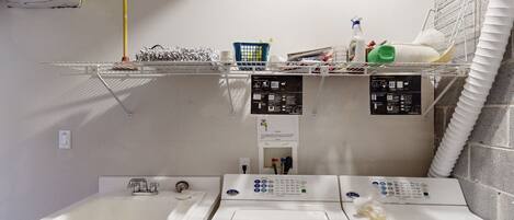 A laundry area with a washing machine, dryer, and a sink. There are shelves above holding various items like bottles and a basket.