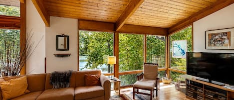 Una sala de estar acogedora con techo de madera, un ventanal grande, un sofá, una silla, un televisor y una estantería.