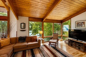 A cozy living room with a wooden ceiling, a large window, a sofa, a chair, a TV, and a bookshelf. - Family Lake House with Deck, Private Dock/Boathouse, Grill, Firepit, & Fast WiFi (Mineral)