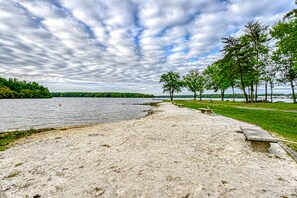 Une plage de sable avec un banc, une zone herbeuse avec des arbres et un lac sous un ciel nuageux.