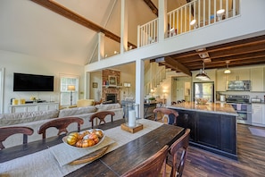 A spacious dining area with a long wooden table, multiple chairs, and a central island with a bowl of fruit. A television is mounted on the wall, and there is a staircase leading to an upper level. - Beautiful lakefront home with fireplace, deck, central AC, & washer/dryer (Mineral)