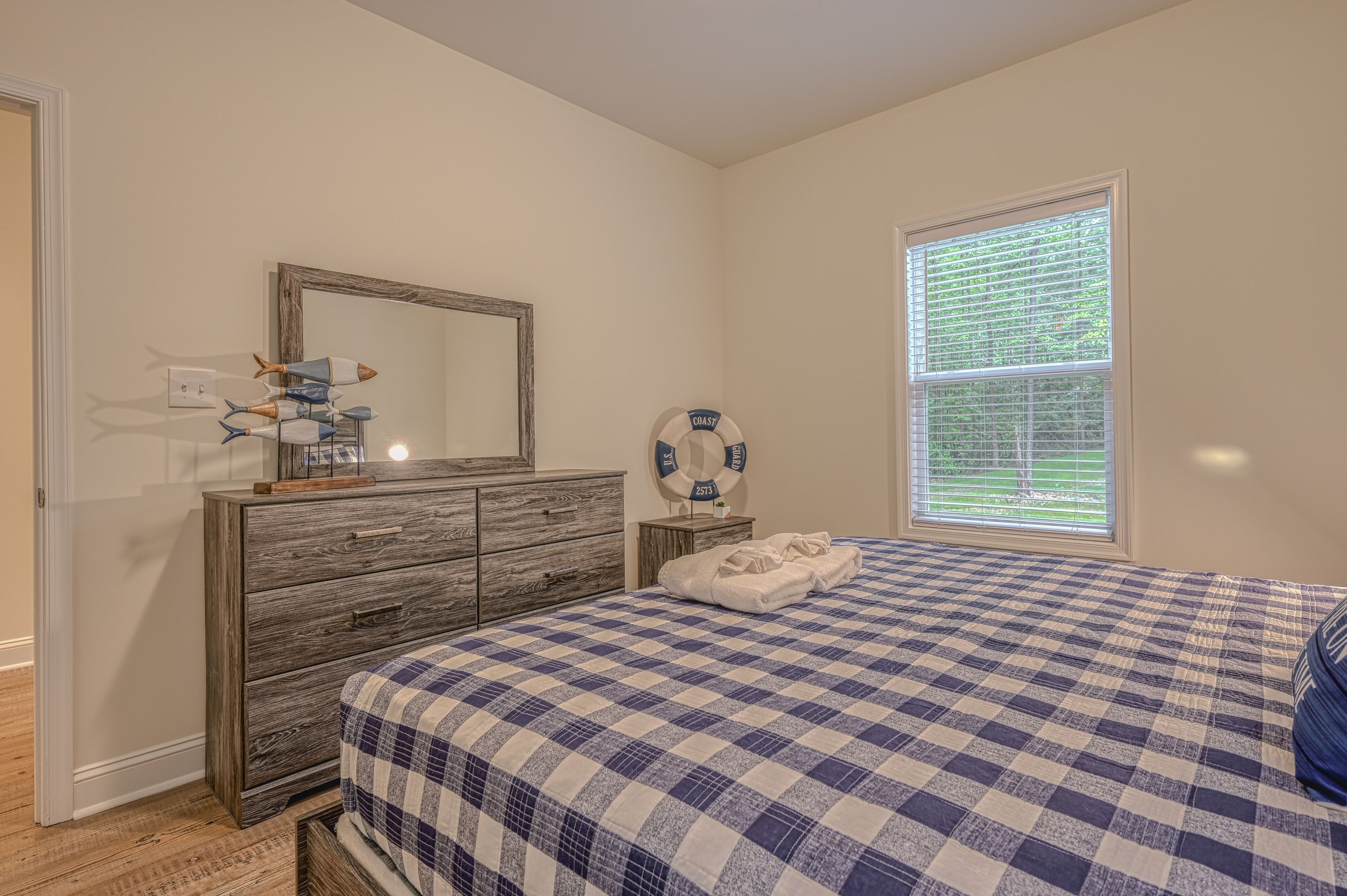 A bedroom with a checkered bedspread, a wooden dresser, a mirror, a life ring decoration, and a window with blinds.