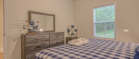A bedroom with a checkered bedspread, a wooden dresser, a mirror, a life ring decoration, and a window with blinds.