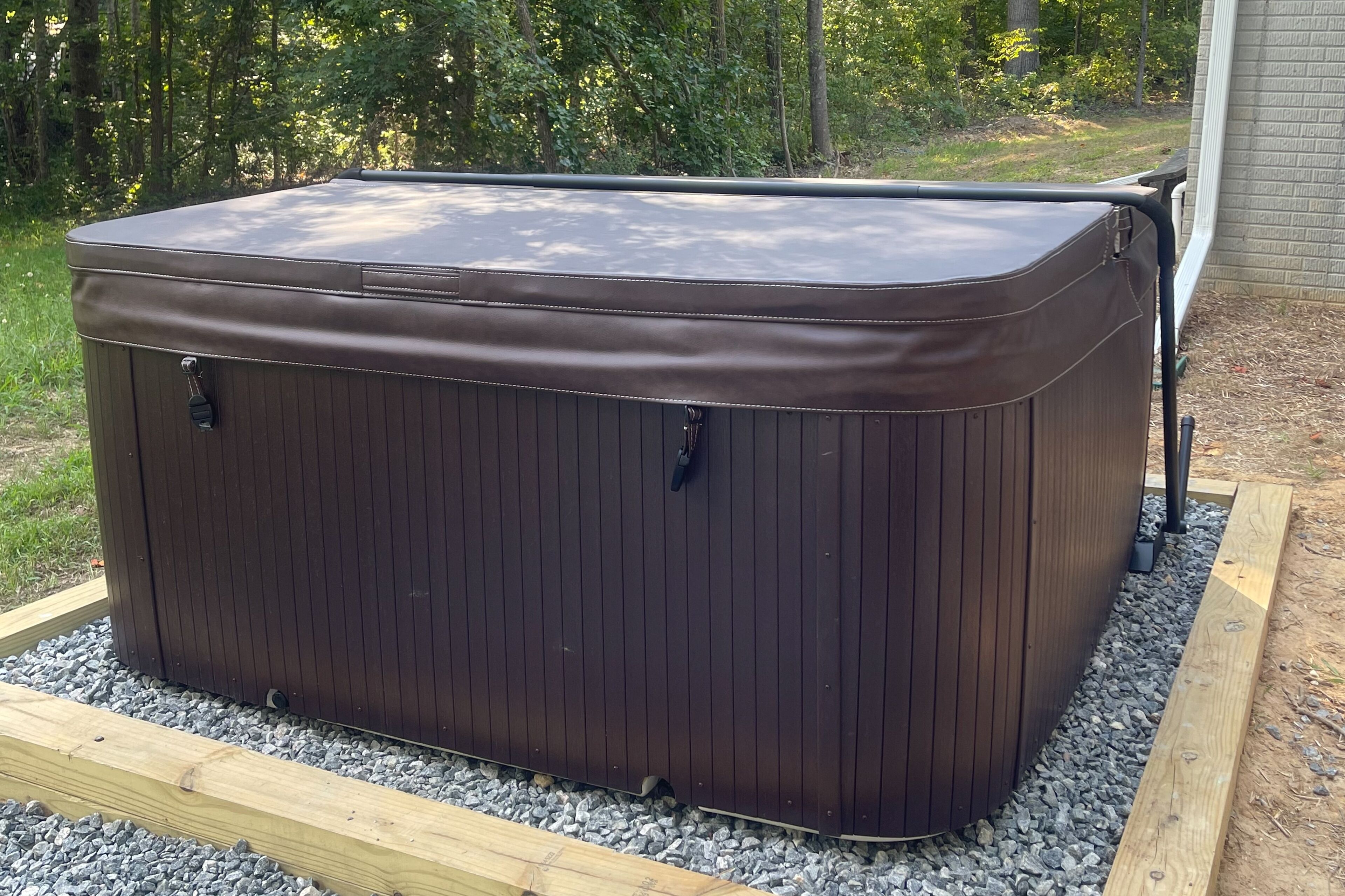 Una bañera de hidromasaje marrón con estructura de madera y patas de metal, ubicada sobre grava y rodeada por una terraza de madera.