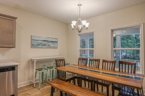 Un comedor con una larga mesa de madera, seis sillas y un banco. Encima de la mesa, un candelabro; en la pared, un cuadro, y una ventana con vistas a árboles.